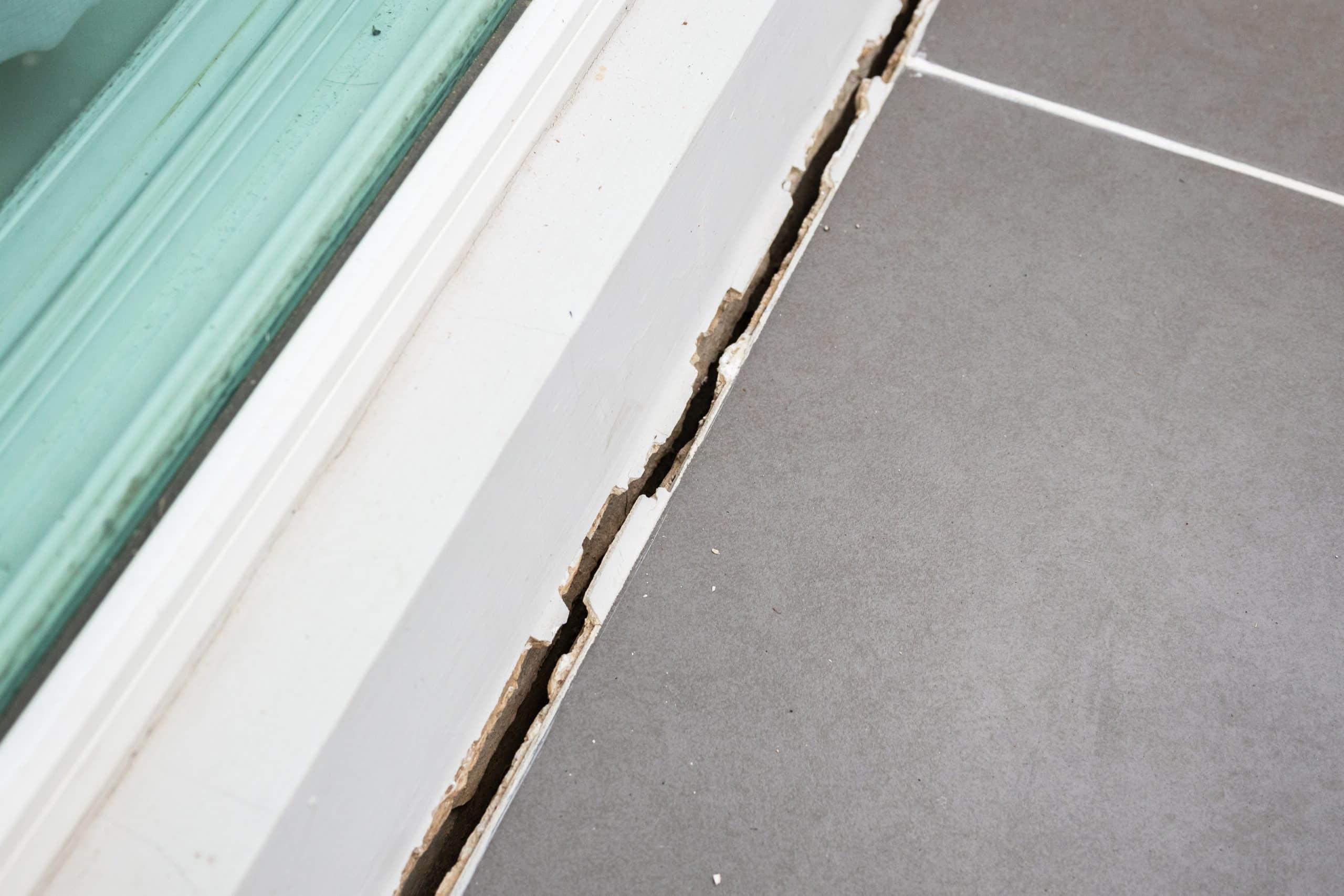 Damaged silicone seal and widening gap between bathroom floor tiles and window frame requiring repair.
