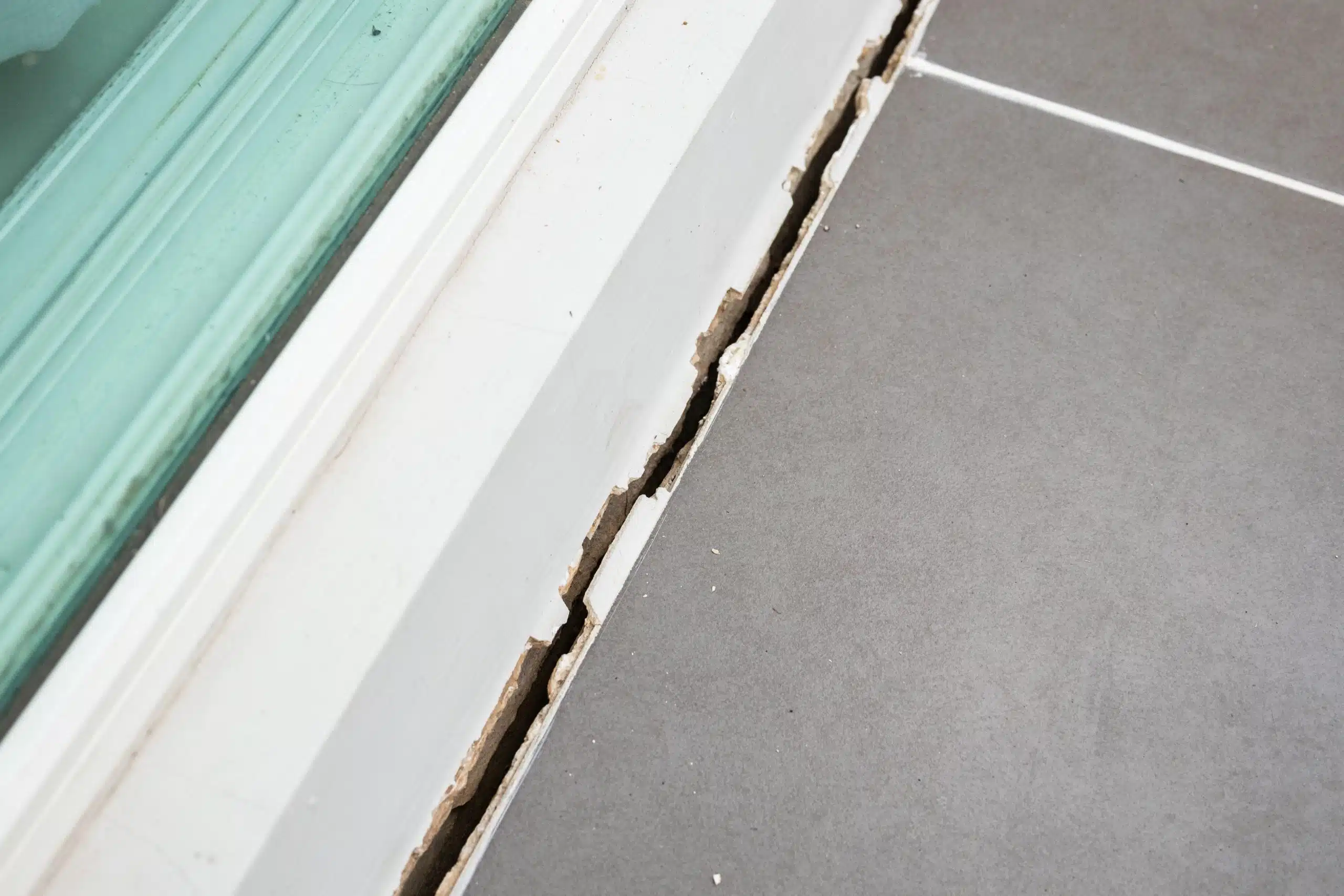 Damaged silicone seal and widening gap between bathroom floor tiles and window frame requiring repair.