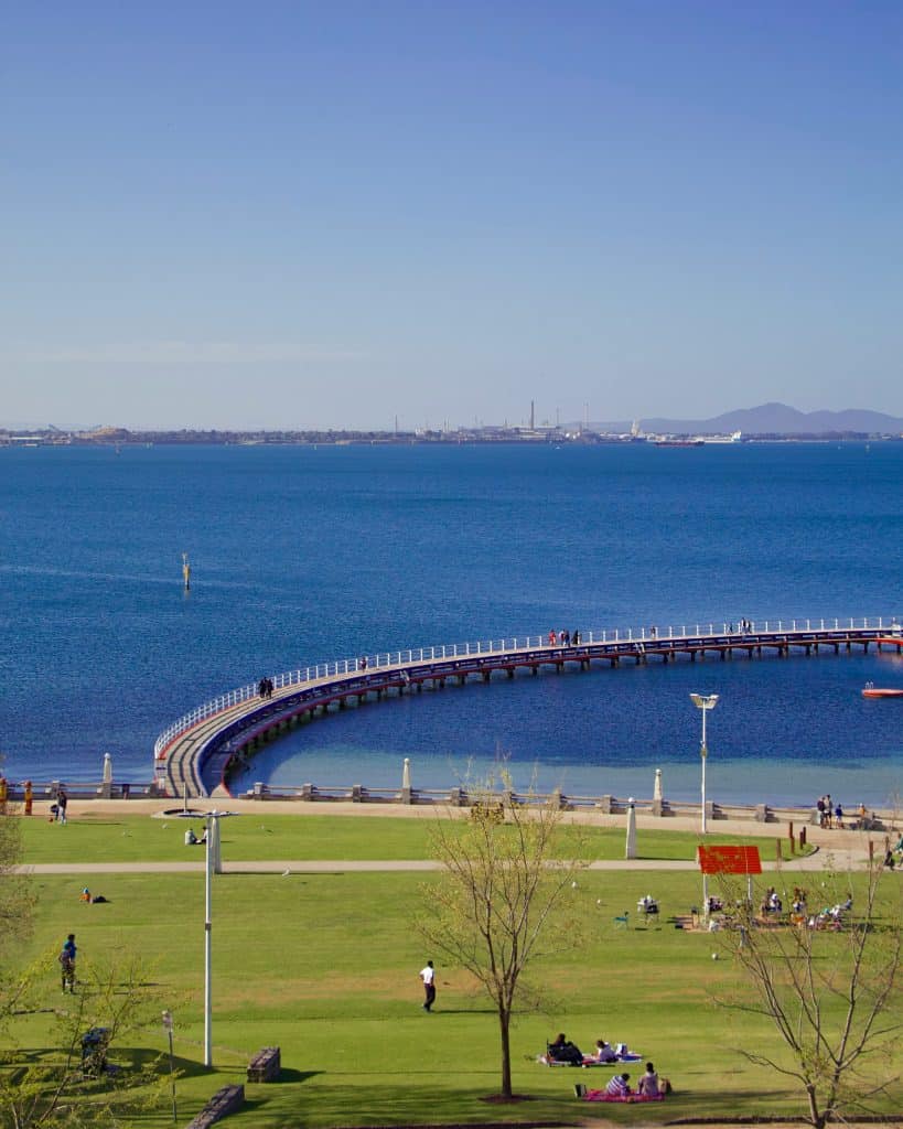 Curved waterfront pier extending into the bay beside a grassy foreshore park with people walking and relaxing.