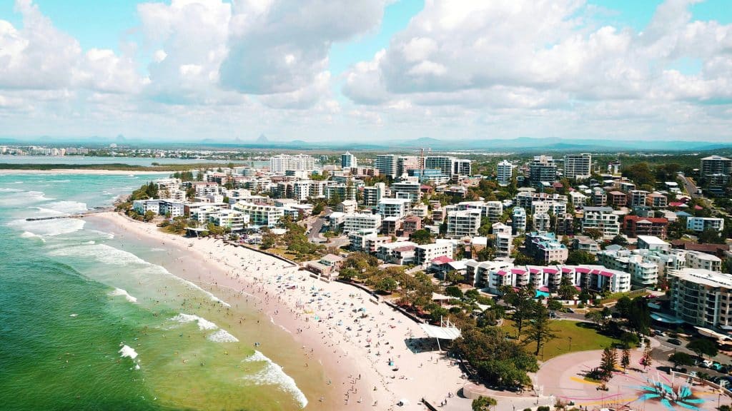 Aerial view of Sunshine Coast beachfront with apartment buildings, sandy beach, ocean waves and people along the shoreline.